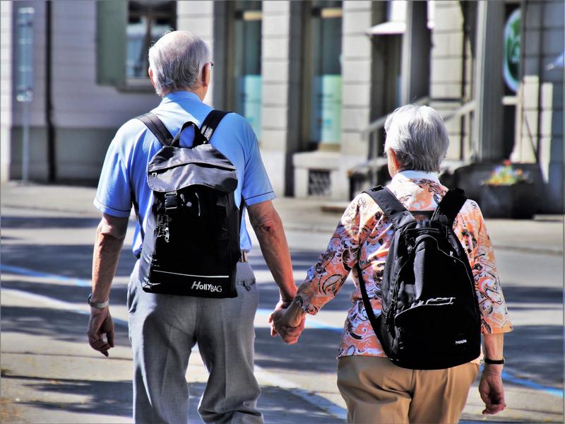 Ein älterer Herr und eine ältere Dame mit Rucksäcken halten sich an den Händen.